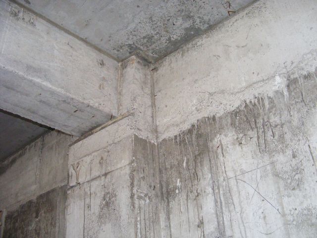Colț de structură din beton aparent cu grinzi, planșeu și urme de scurgeri pe suprafață.
