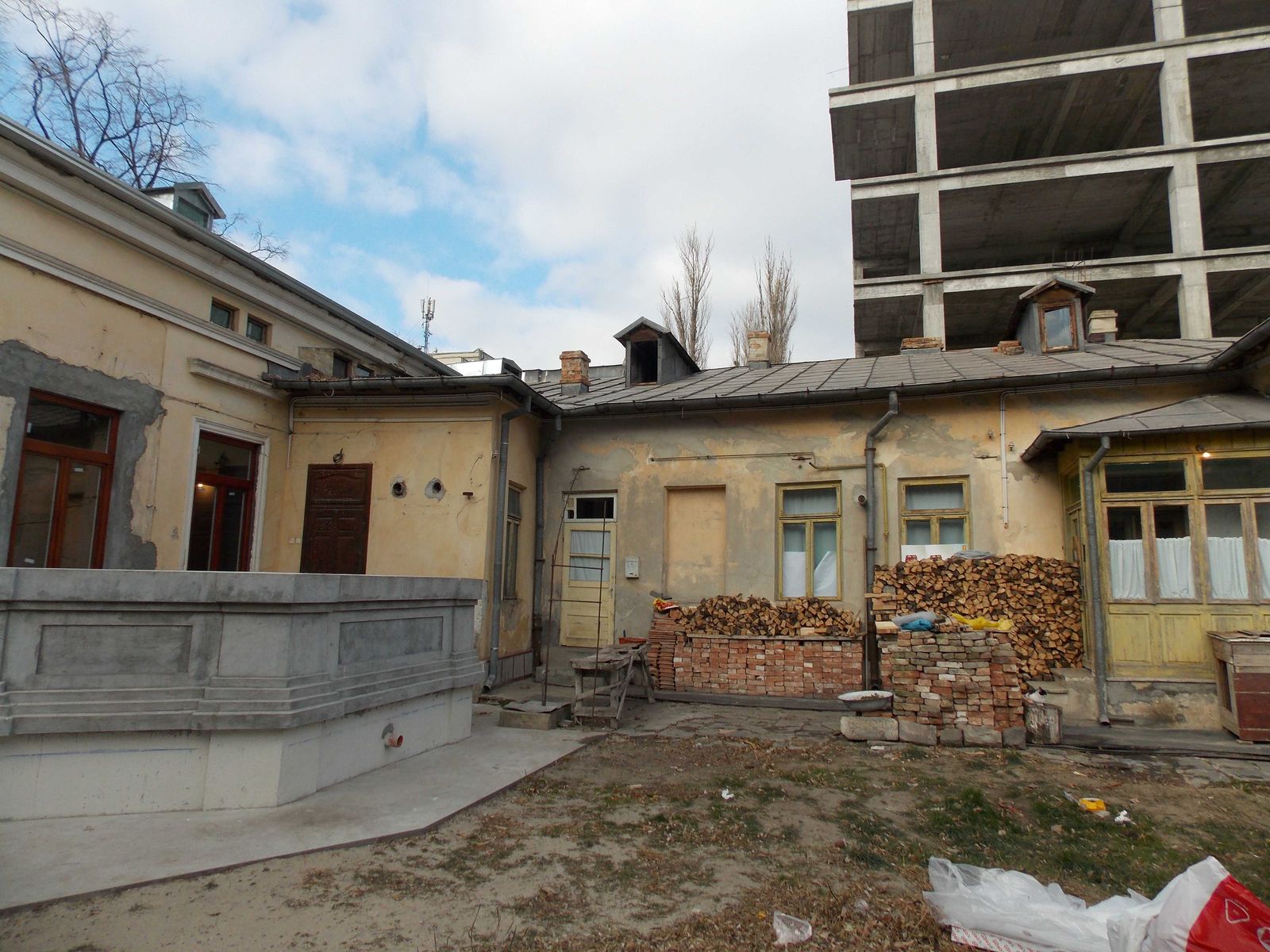 Fațadă joasă de zidărie tencuită, cu acoperiș din tablă, și o clădire vecină în schelet de beton armat în spatele ei.
