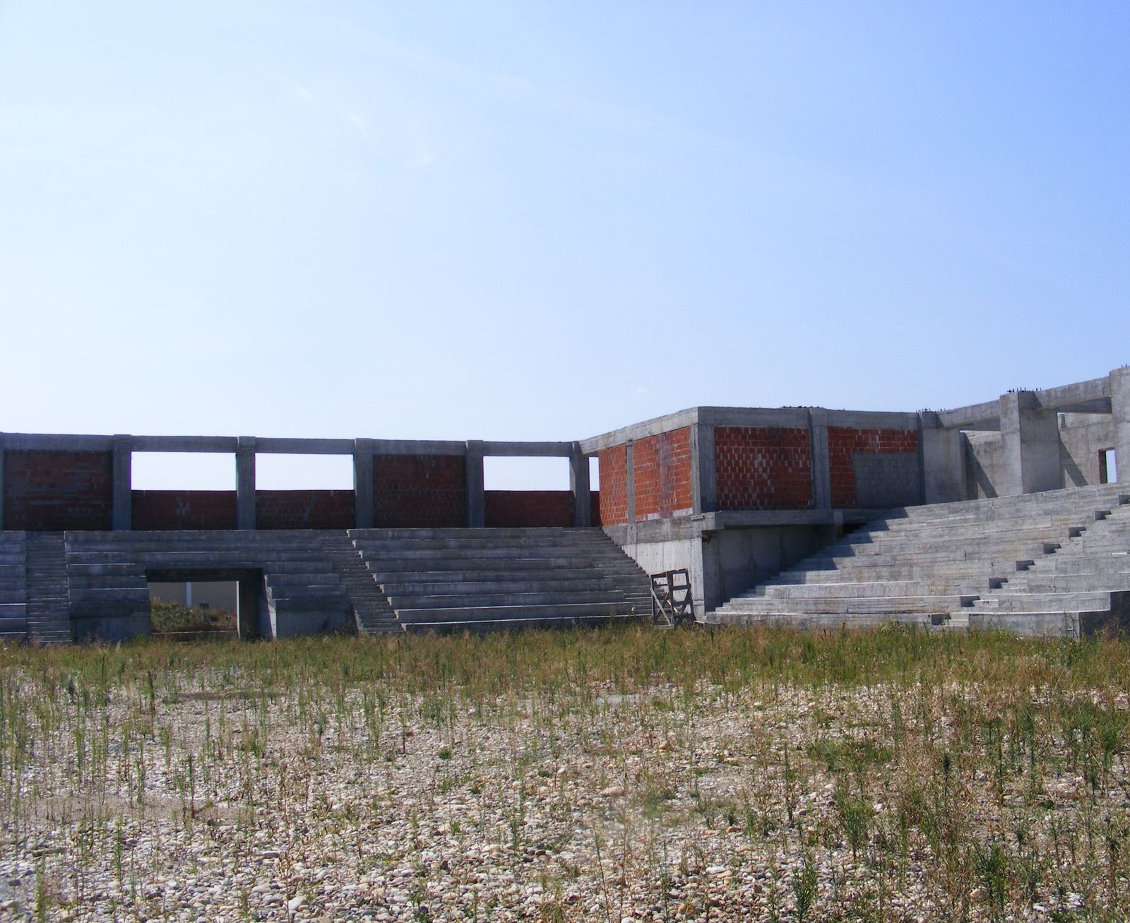 Structura de beton armat a sălii de sport, cu tribune, văzută dintr-o pajiște, fără acoperiș